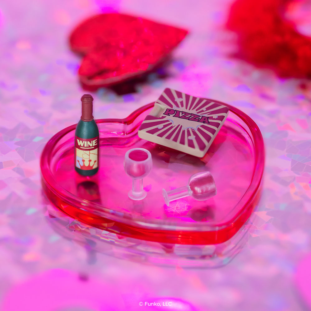This photo shows a heart-shaped red-hued plastic tray holding a wine bottle accessory, two wine glass accessories, and a pizza box accessory. Glittery hearts are in the background.